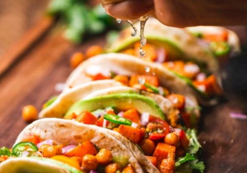 The image shows a hand squeezing a lime over a row of colorful tacos filled with chickpeas, avocado, and vegetables on a wooden board.