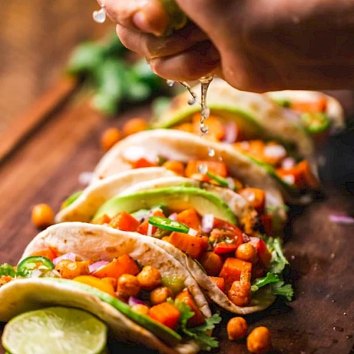 The image shows a hand squeezing a lime over a row of colorful tacos filled with chickpeas, avocado, and vegetables on a wooden board.