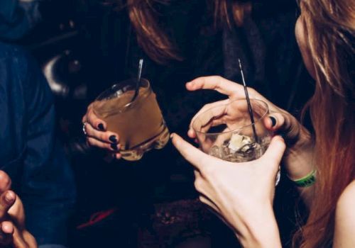 A group of people are holding drinks and chatting in a dimly lit setting. Two of them are visible, and one person appears to be laughing.