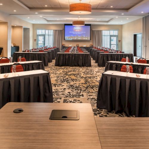 A conference room with tables arranged in a U-shape, red chairs, and two large screens displaying a cityscape and the text 