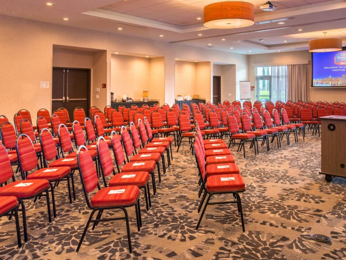 The image shows a conference room with red chairs arranged in rows facing a projector screen displaying a presentation.