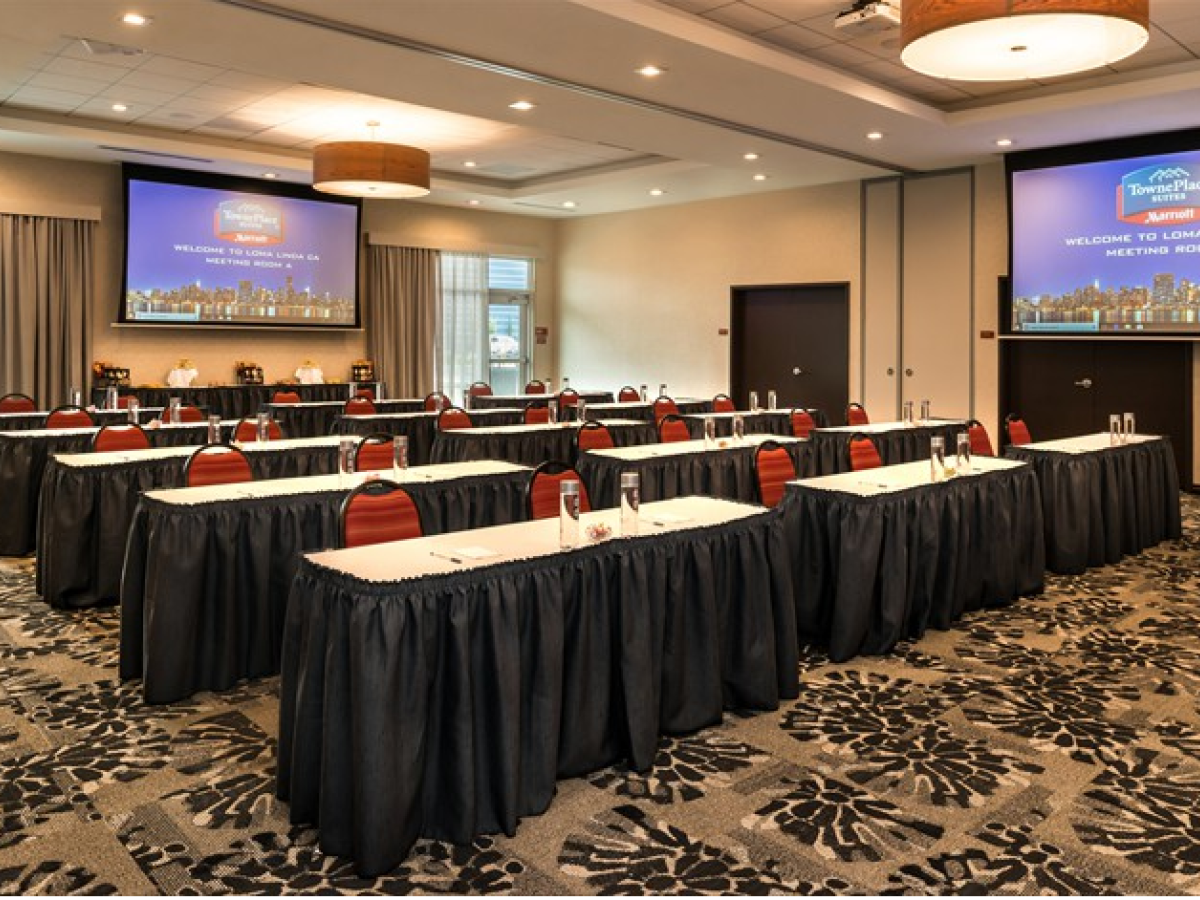 This image shows a conference room set up with long tables, red chairs, water glasses, notebooks, and two large projection screens at the front.