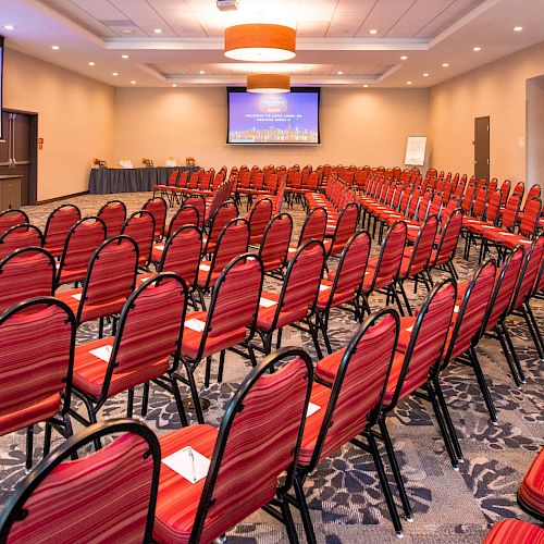 The image shows an empty conference room with red chairs arranged in rows facing a presentation screen. There are two screens displaying content.