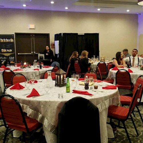 The image shows an indoor event with several round tables covered in white cloths and red napkins, with attendees seated and conversing.