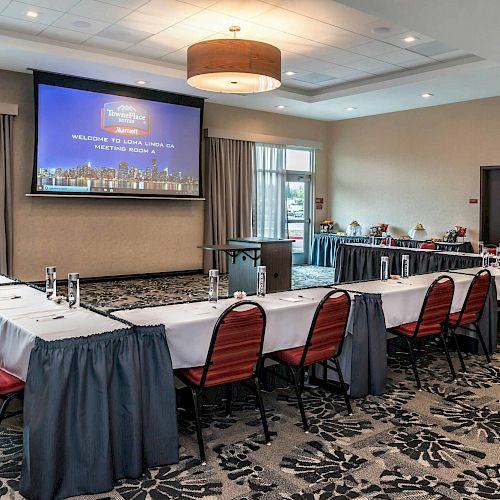 The image shows a conference room set up with U-shaped tables, red chairs, a podium, and a large screen displaying a welcome message.