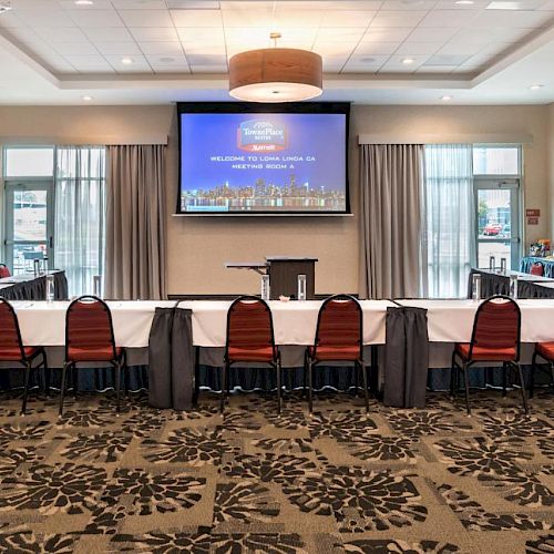 A conference room with U-shaped table setup, red chairs, large screen displaying presentation, podium in the center, and decorative carpet flooring.