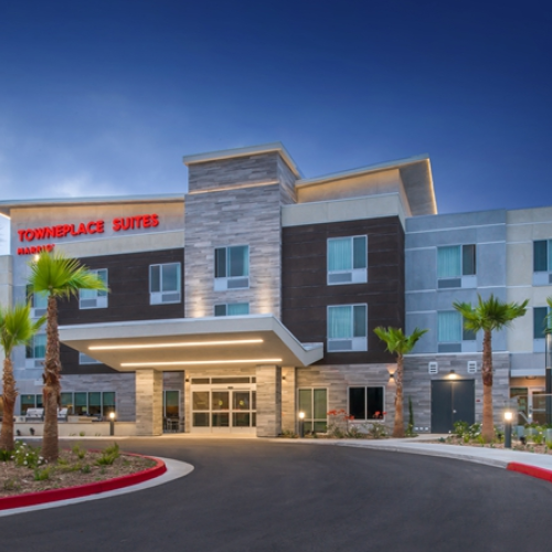 The image shows the exterior of a TownePlace Suites by Marriott hotel with a modern design, palm trees, and a welcoming entrance.