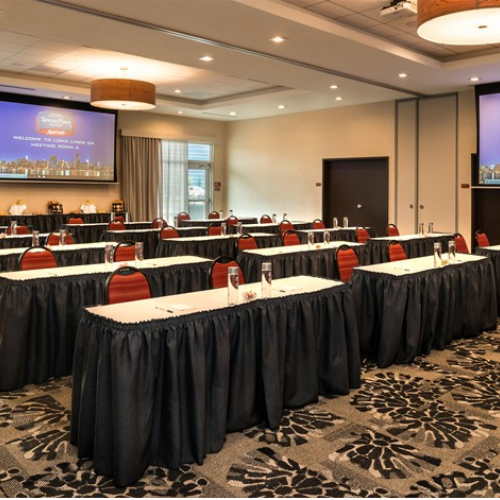 The image shows a conference room set up with red chairs, tables with black skirts, and two large screens displaying a welcome message.