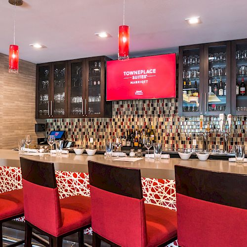 This image shows a modern bar area with red cushioned stools, a countertop, and glass cabinets behind. A screen displays 