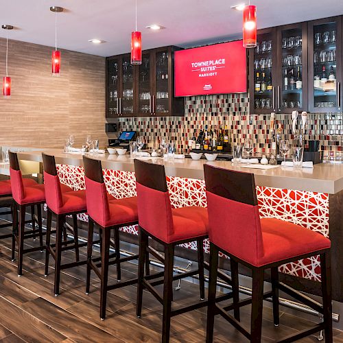 A modern bar with red chairs, pendant lights, a tiled backsplash, and a countertop, finished with a display of bottles and glasses.