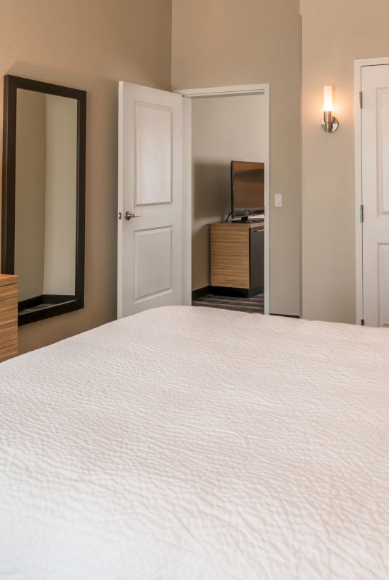 A neatly organized bedroom features a bed with white linens, a TV on a dresser, a wall mirror, and doors leading to other rooms.