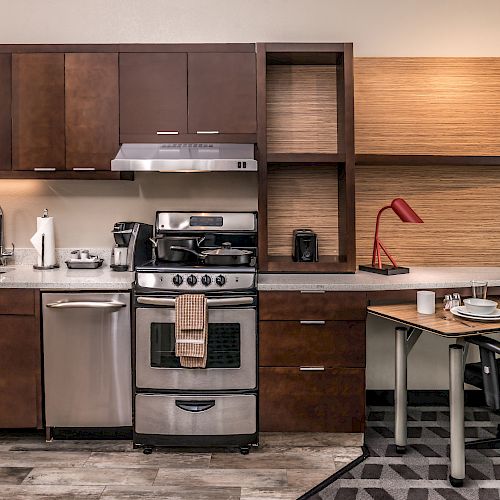 A modern kitchen setup with wooden cabinets, a stove, a microwave, a sink, and a desk with a lamp and chair on the right side, ending the sentence.