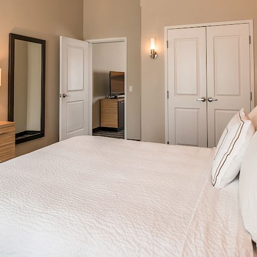 This image shows a neatly made bed in a modern bedroom with a TV, dresser, mirror, and closed double doors, creating a cozy ambiance.