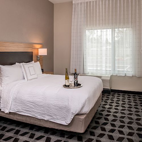 A neatly arranged hotel room with a queen-sized bed, two bedside tables, a TV, and a window; wine bottles and glasses on the bed.