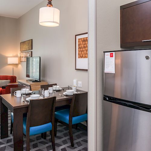 The image shows a modern living space with a dining table, chairs, a stainless-steel refrigerator, and a cozy living area with a red chair and TV.