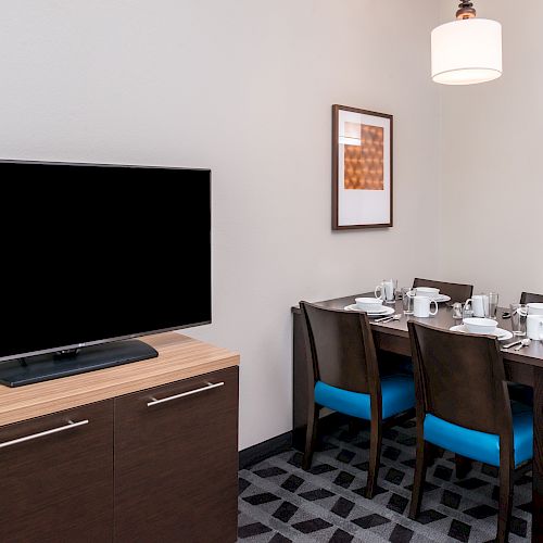 A modern dining area with four chairs, a set dining table, a wall-mounted TV, a pendant light, artwork, and part of a kitchenette in the background.