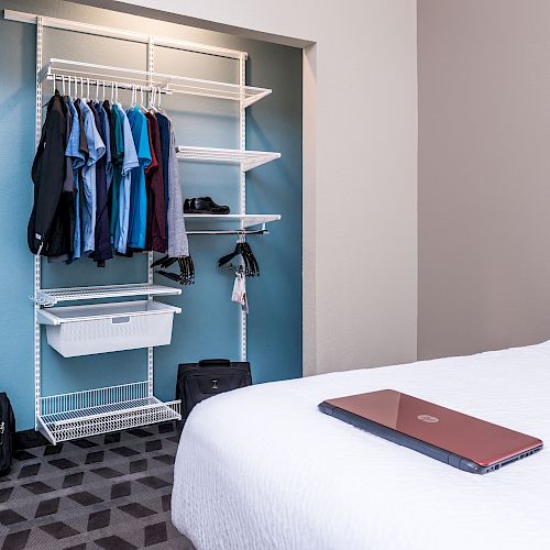 A neatly organized room with a closet holding shirts, a laptop on a bed, and backpacks on the ground. The space feels clean and efficient.