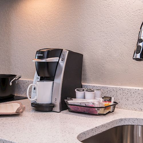 The image shows a kitchen countertop with a coffee maker, coffee pods, a pot, a folded towel, and a sink with a curved faucet, ending the sentence.